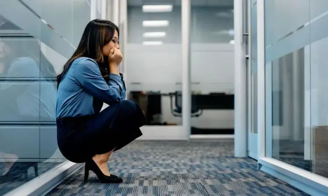Frau kauert im Büroflur, Hände vor dem Mund, die Augen geschlossen. Sie scheint von **Angst vor der Arbeit** überwältigt zu sein.