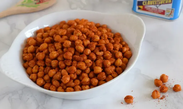 Un tazón blanco lleno de garbanzos fritos crujientes y especiados. Una cuchara de madera decorada y una lata de especias están al fondo.