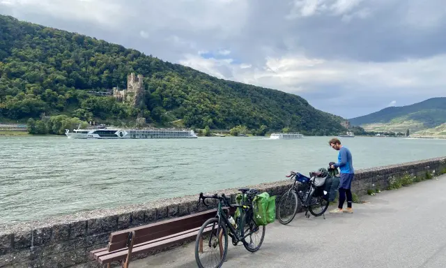 Mann packt Gepäck für eine Fahrradtour am Rhein. Im Hintergrund Burg und Fluss.