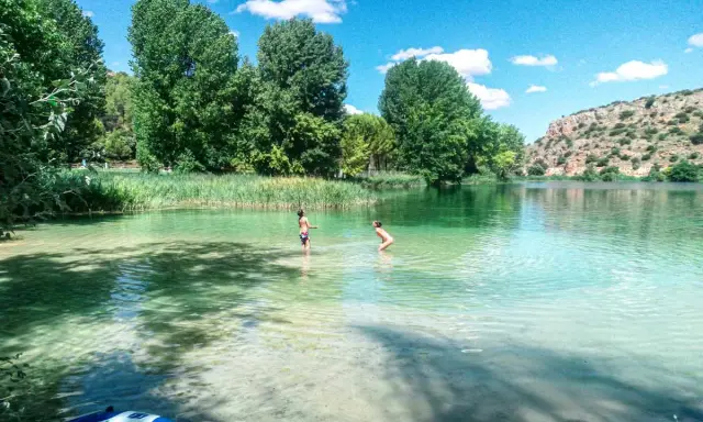 Dos niños juegan en las cristalinas aguas de las Lagunas de Ruidera, rodeados de vegetación y un paisaje montañoso.