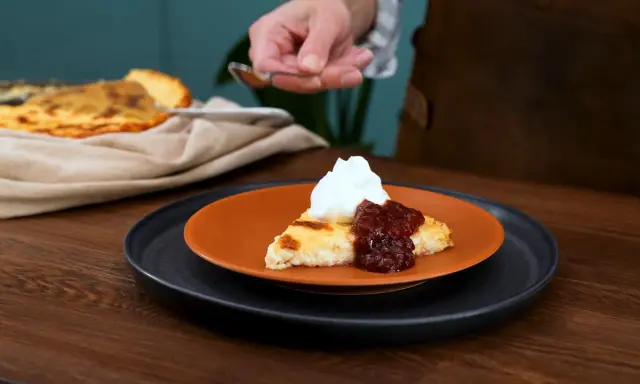 En bit krämig ostkaka med keso, toppad med vispgrädde och sylt. Serveras på en orange tallrik.