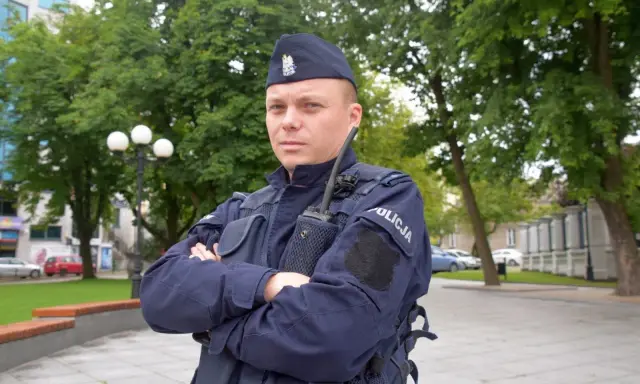 Policjant w granatowym mundurze, w sprawie umundurowania policjantów, stoi na tle zieleni.