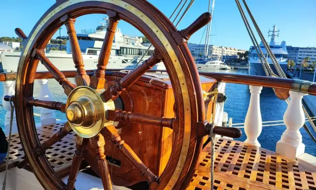Roue de gouvernail en bois et laiton sur le pont d'un bateau. En arrière-plan, des yachts modernes et des bâtiments dans un port ensoleillé.