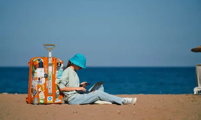 Dziewczyna w niebieskim kapeluszu siedzi na plaży z walizką, laptopem i telefonem, planując, jak tanio podróżować.