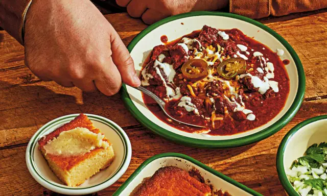 En hand tar en sked ur en skål med mustig texas chili, toppad med jalapeños och gräddfil. Bredvid ligger gyllene majsbröd.