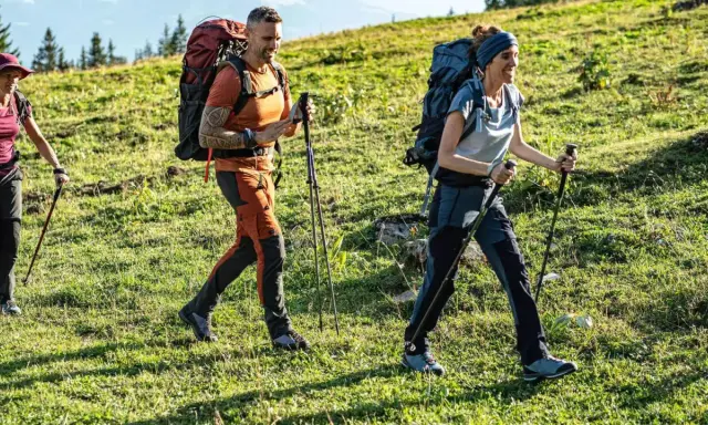 Trzy osoby wędrują po zielonym zboczu. Mężczyzna i kobieta w wygodnych, sportowych spodniach w góry, z plecakami i kijkami trekkingowymi.