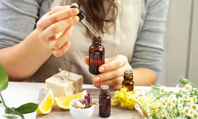 Une personne prépare un parfum huile essentielle, ajoutant une goutte d'ambre dans un flacon. Des fleurs, des citrons et du savon sont disposés autour.