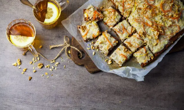 En skiva vegansk morotskaka med glasyr och nötter, serverad med två koppar te med citron.