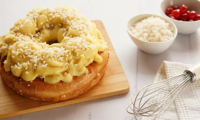Rosca de Pascua decorada con crema pastelera y azúcar granulada, lista para disfrutar. A su lado, cuencos con cerezas y ralladura, y un batidor.