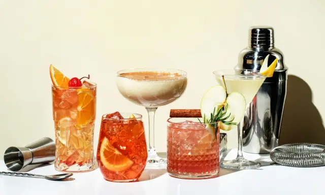 Eine Auswahl an fruchtigen Cocktails, garniert mit Orangen, Äpfeln und Kirschen, neben einem Cocktail-Shaker.