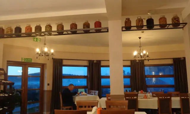Wnętrze restauracji z oknami wychodzącymi na góry, stoły nakryte obrusami, a na półce nad nimi słoiki z przetworami.