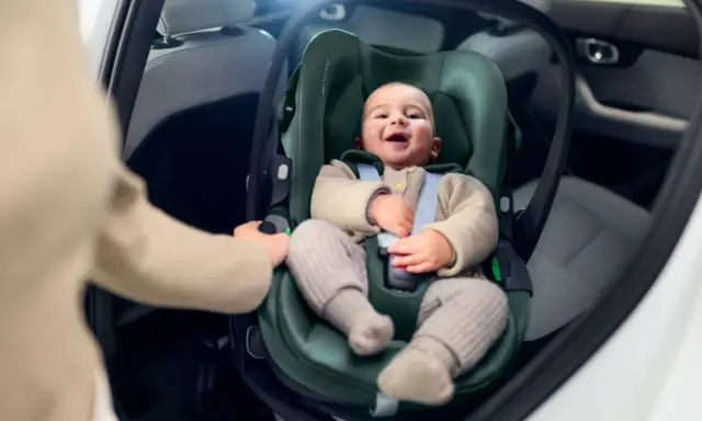 Uśmiechnięte dziecko w foteliku samochodowym. Ręka rodzica pokazuje, jak wypiąć fotelik z bazy isofix.
