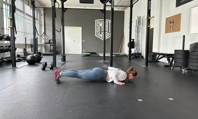 Frau macht Liegestütze im Fitnessstudio, ein Teil ihres plyometrischen Trainings. Hanteln und Medizinbälle liegen bereit.