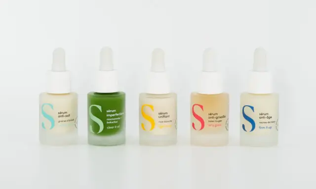 Cinq flacons de sérum et crème hydratante aux couleurs vives, chacun avec une étiquette indiquant son usage : anti-soif, imperfections, unifiant, anti-grisaille, anti-âge.