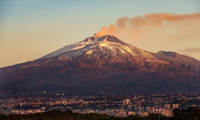 Etna fumante, con la cima innevata che brilla al tramonto, domina una città siciliana.