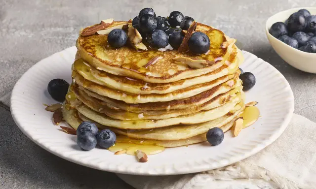 Stapel fluffiger High Protein Pancakes mit Blaubeeren und Mandelsplittern, beträufelt mit Honig.