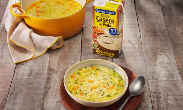 Un tazón humeante de sopa de pollo casera, con fideos finos y trozos de zanahoria, junto a una olla amarilla y un cartón de caldo de pollo.
