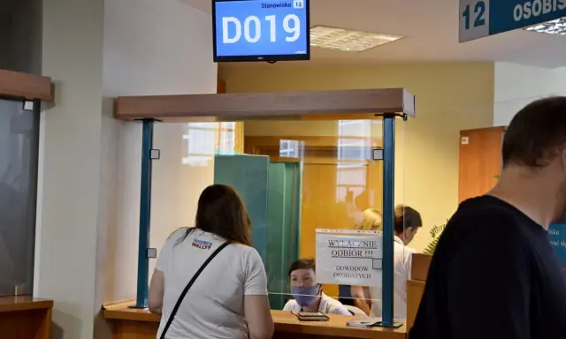 Kobieta czeka na odbiór dowodu rejestracyjnego. Na ekranie wyświetla się numer stanowiska D019.