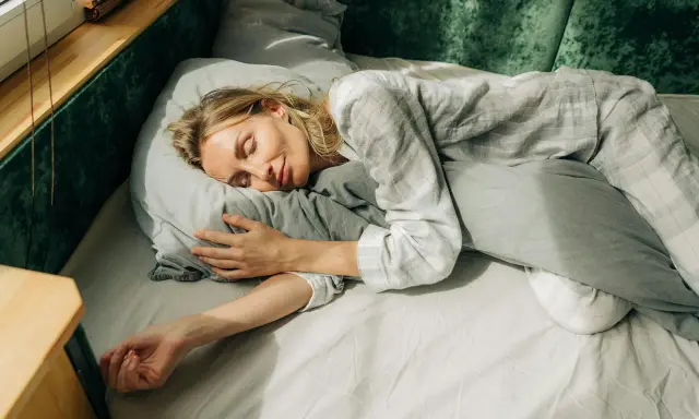 En kvinna sover lugnt i sängen, omfamnande en kudde. Solstrålar faller in genom fönstret och skapar en känsla av guidad avslappning.
