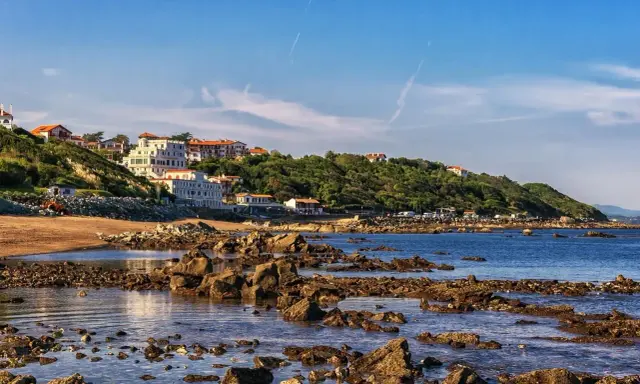 Vue côtière avec des maisons blanches sur une colline verdoyante, une plage de sable et des rochers dans l'eau. Le cadastre zone inondable est visible.