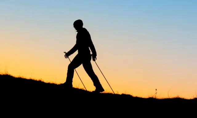 Sylwetka osoby uprawiającej nordic walking na tle zachodzącego słońca. Czy wolisz nordic walking rano czy wieczorem?