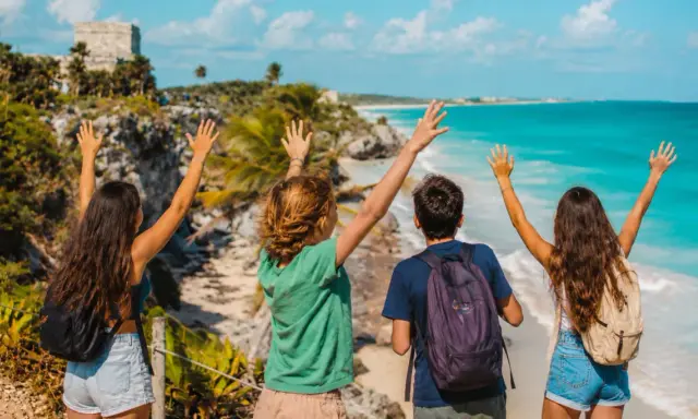 Cuatro jóvenes con mochilas saludan al mar turquesa y las ruinas mayas. ¡Una aventura inolvidable en la Riviera Maya con niños!