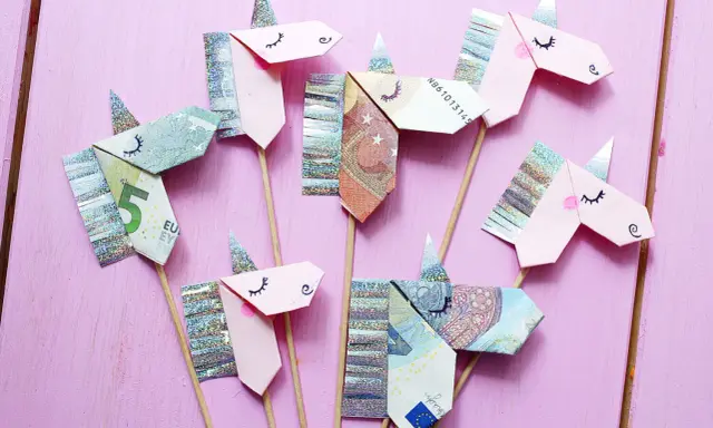 Kreatives Geld falten: Mehrere Einhorn-Figuren aus Geldscheinen und Glitzerpapier auf Holzstäbchen vor rosafarbenem Hintergrund.