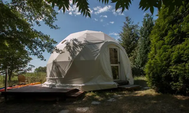 Biały namiot kopułowy, idealny na glamping noclegi, otoczony zielenią. Drewniany taras z meblami zachęca do relaksu.