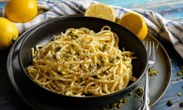 Eine Schüssel mit Zitronen Parmesan Pasta, garniert mit gehackten Pistazien und schwarzem Pfeffer. Frische Zitronen liegen daneben.
