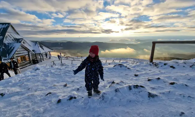 Radosne bieszczady z dziećmi. Mała dziewczynka w czerwonej czapce cieszy się śniegiem na szczycie góry, w tle widać schronisko i piękne zachmurzone niebo.