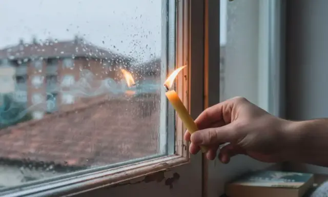 Una vela encendida cerca de una ventana con gotas de lluvia. El humo de la vela se arremolina, y parece que entra aire por la ventana, reflejando edificios en el exterior.