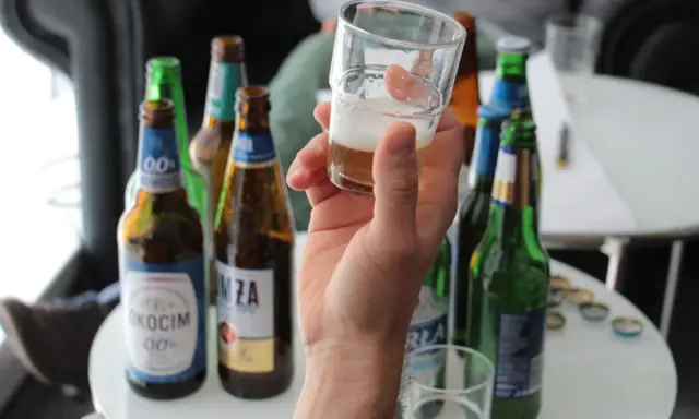 Ręka trzyma kieliszek z piwem bezalkoholowym. W tle kilka butelek piwa, idealne na przerwę w pracy.