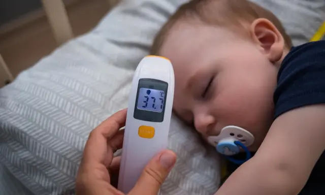 Elektroniczny termometr pokazuje 37.7°C, mierząc temperaturę niemowlaka śpiącego ze smoczkiem.