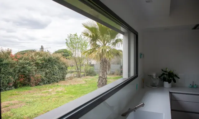 Vista desde una cocina moderna con **ventanas automáticas** que enmarcan un jardín con palmera y vegetación frondosa.