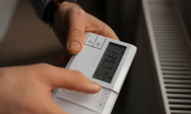 Dłoń naciska przycisk na białym sterowniku, by ustawić temperaturę na grzejniku.