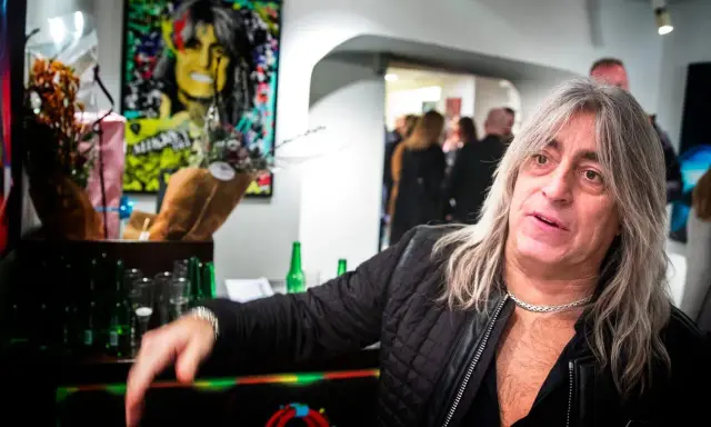 Mikkey Dee son, en man med långt grått hår och en silverfärgad kedja, poserar vid ett bord med blommor och flaskor.