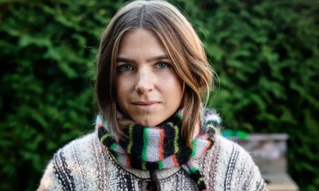 Elin Berlin med blå ögon och fräknar, klädd i en stickad tröja och randig halsduk, står framför en grön häck.