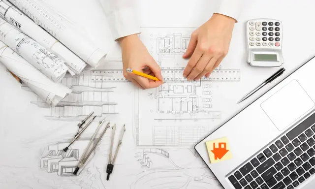 Manos de arquitecto dibujando planos, con calculadora, laptop y bocetos. El significado de "as built" se refleja en la precisión del diseño.