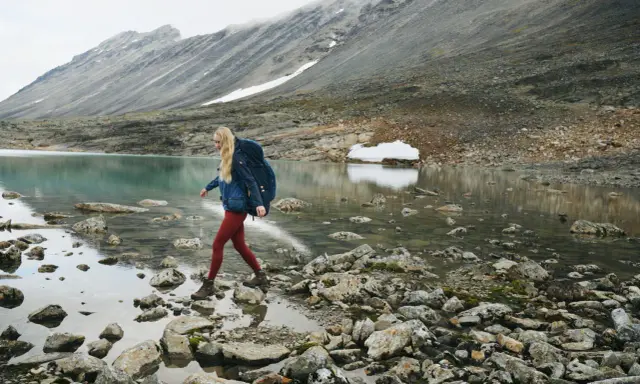 Kobieta z plecakiem przemierza kamieniste brzegi górskiego jeziora. W tle skaliste zbocza, idealne dla skandynawskie marki outdoorowe.