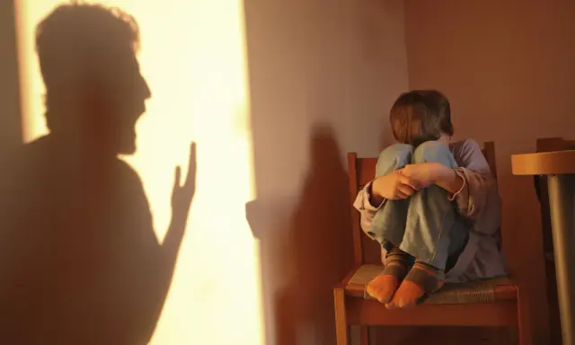 Cień krzyczącej postaci rzuca się na ścianę, podczas gdy dziecko skulone na krześle odczuwa skutki bicia dzieci w dorosłym życiu.