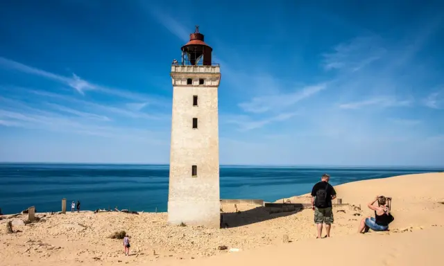 Ett vitt utsiktstorn i Danmark reser sig mot en blå himmel. Sanddyner och hav omger tornet.