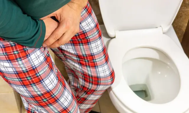 Mężczyzna w piżamie z kratę trzyma się za krocze, siedząc obok toalety. Czy zwapnienie prostaty to rak?