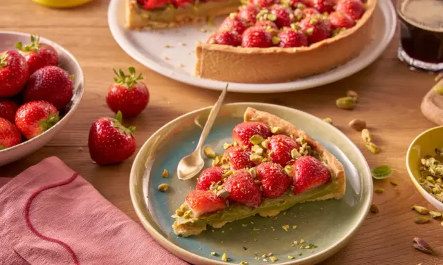 Kawałek tarty z kremem pistacjowym i truskawkami, posypany pistacjami. Obok cała tarta, miseczka z truskawkami i filiżanka kawy.