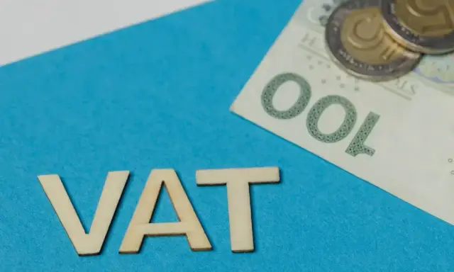 Drewniane litery VAT na niebieskim tle, obok banknot 100 zł i monety. Symbolizuje zwolnienie z VAT.