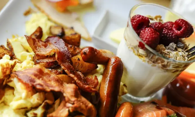Herzhafte Ideen zum Brunch: Rührei, Speck, Würstchen und Kartoffeln neben einem Glas Joghurt mit Beeren und Müsli.