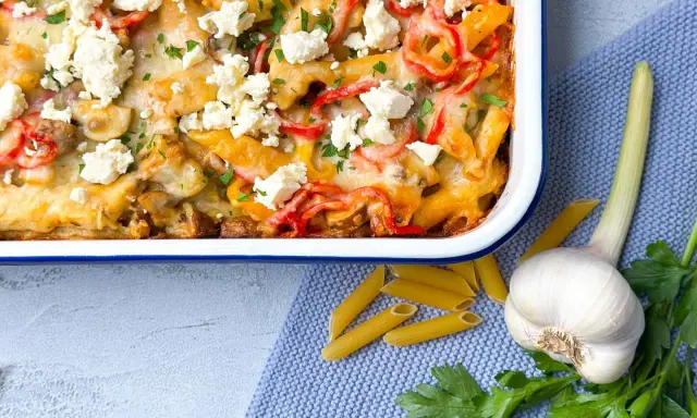 Herzhafter Gyros Auflauf mit Penne, Paprika und Feta, garniert mit Petersilie. Daneben liegen Penne, Knoblauch und frische Kräuter.