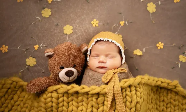 Bambino addormentato con orsacchiotto, avvolto in una coperta gialla. Un'idea per foto neonati fai da te con decorazioni floreali.