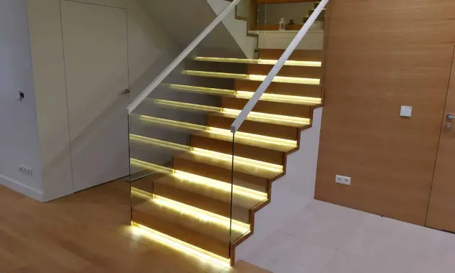 Nowoczesne schody dywanowe na beton z podświetleniem LED. Szklana balustrada i drewniane stopnie tworzą elegancką całość.