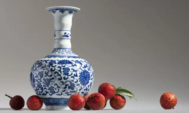 Un vase Satsuma bleu et blanc orné de motifs floraux, entouré de litchis rouges. Comment reconnaître un vase Satsuma ? Observez les détails.