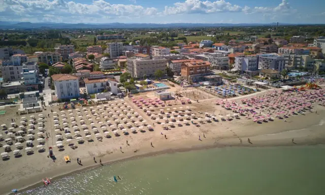 Vista aerea di una spiaggia affollata con ombrelloni rosa e bianchi, hotel e case nel centro storico di Torre Pedrera.